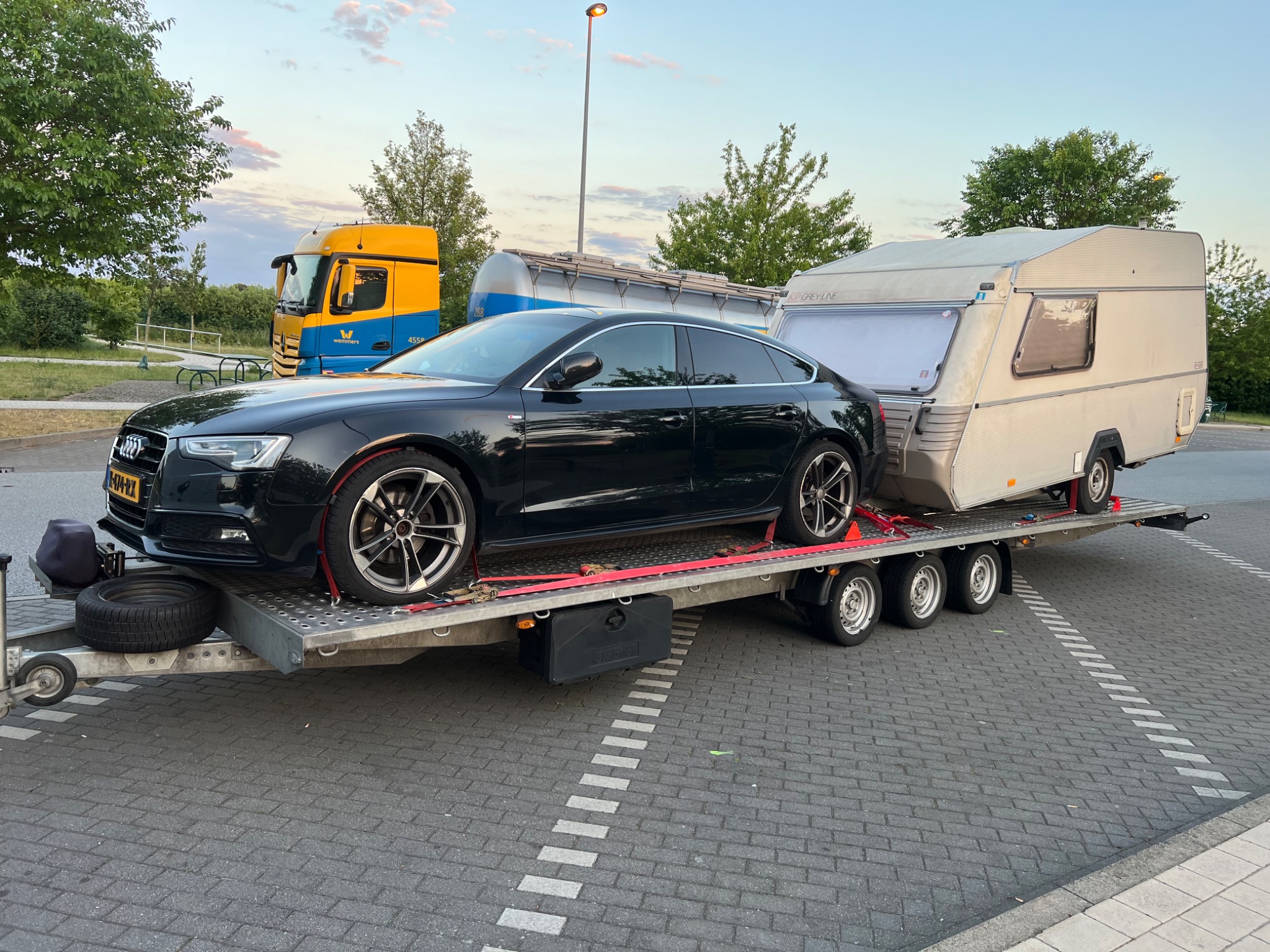 Czarny samochód osobowy Audi A5 i przyczepa kempingowa na lawecie, zabezpieczone czerwonymi pasami transportowymi, w tle widoczny żółto-niebieski samochód ciężarowy.