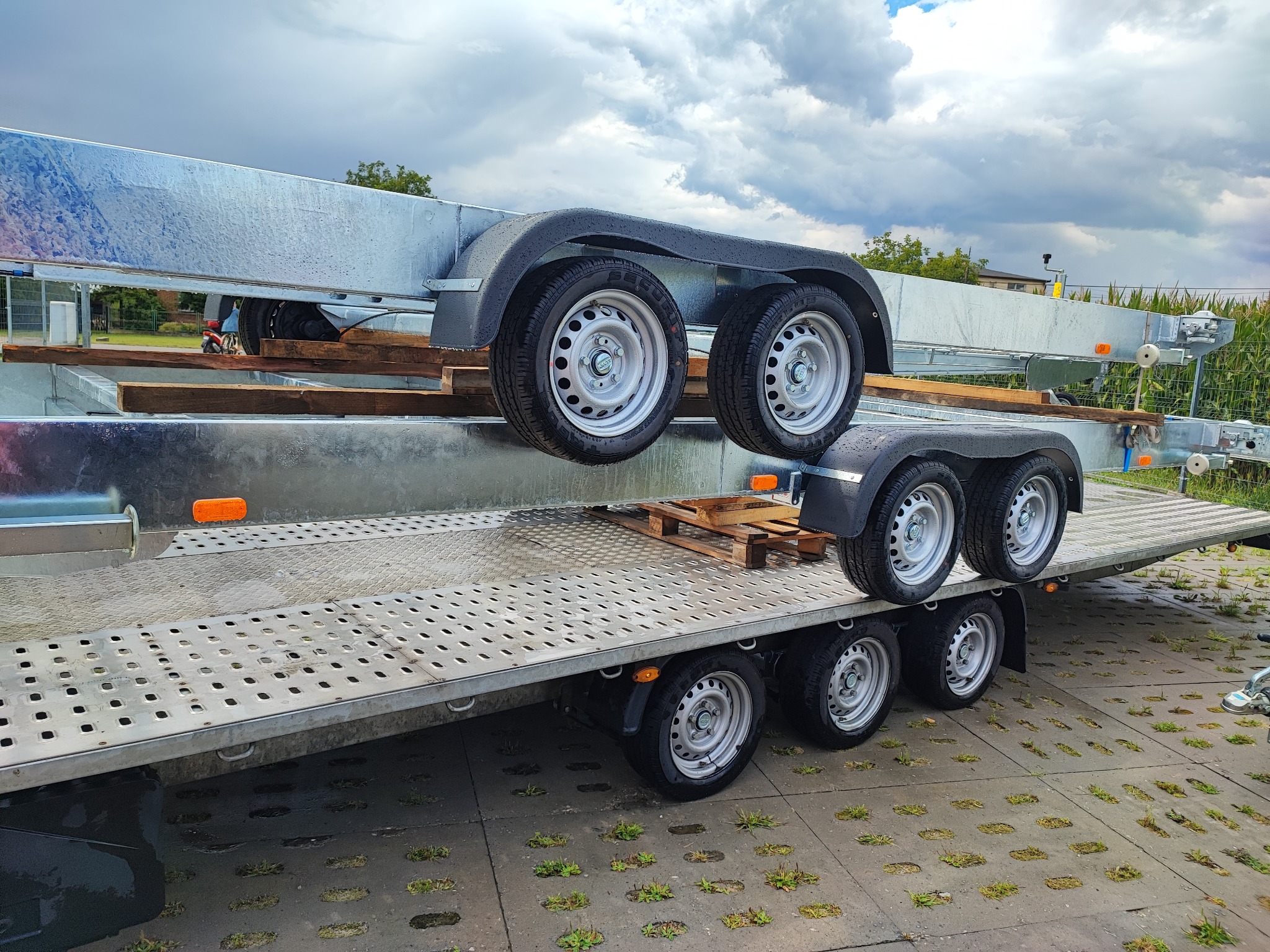 Dwa nowe, ocynkowane przyczepy samochodowe piętrowo ułożone na betonowym placu z trawą, widoczne detale kół i konstrukcji.