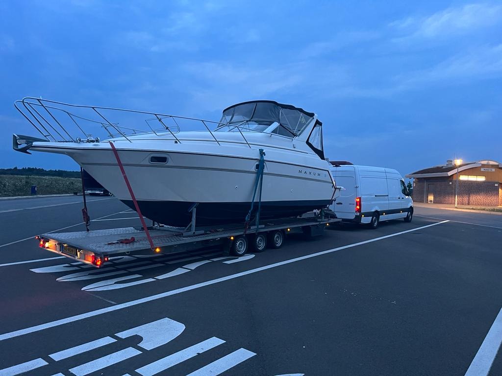 Biała łódź motorowa marki Maxum zabezpieczona czerwonymi pasami na niskopodwoziowej przyczepie, holowana przez białego busa na parkingu w pochmurny dzień.