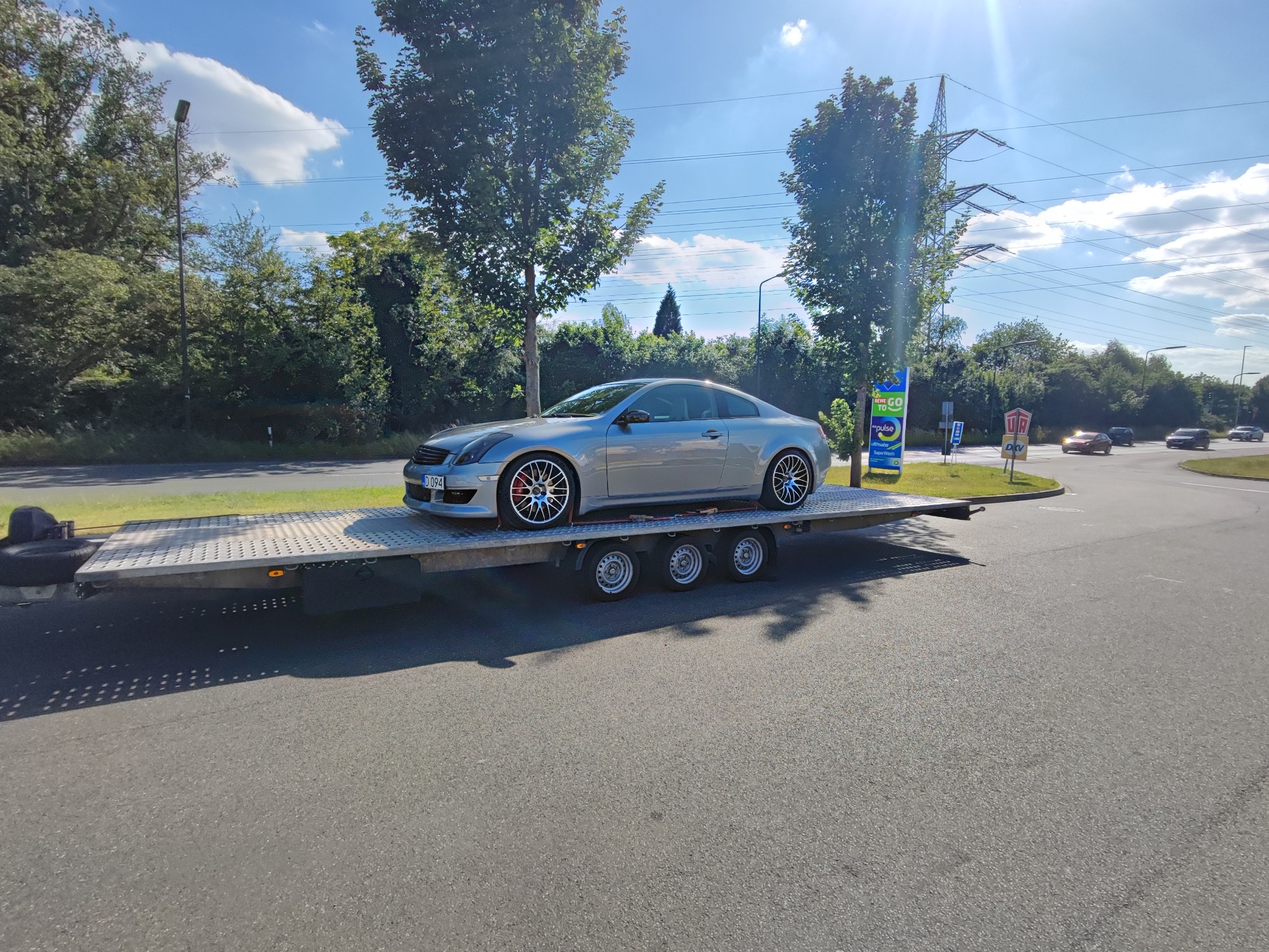 Srebrny sportowy samochód marki Infiniti na niskopodwoziowej przyczepie transportowej, widok z boku na tle zieleni i błękitnego nieba z chmurami.