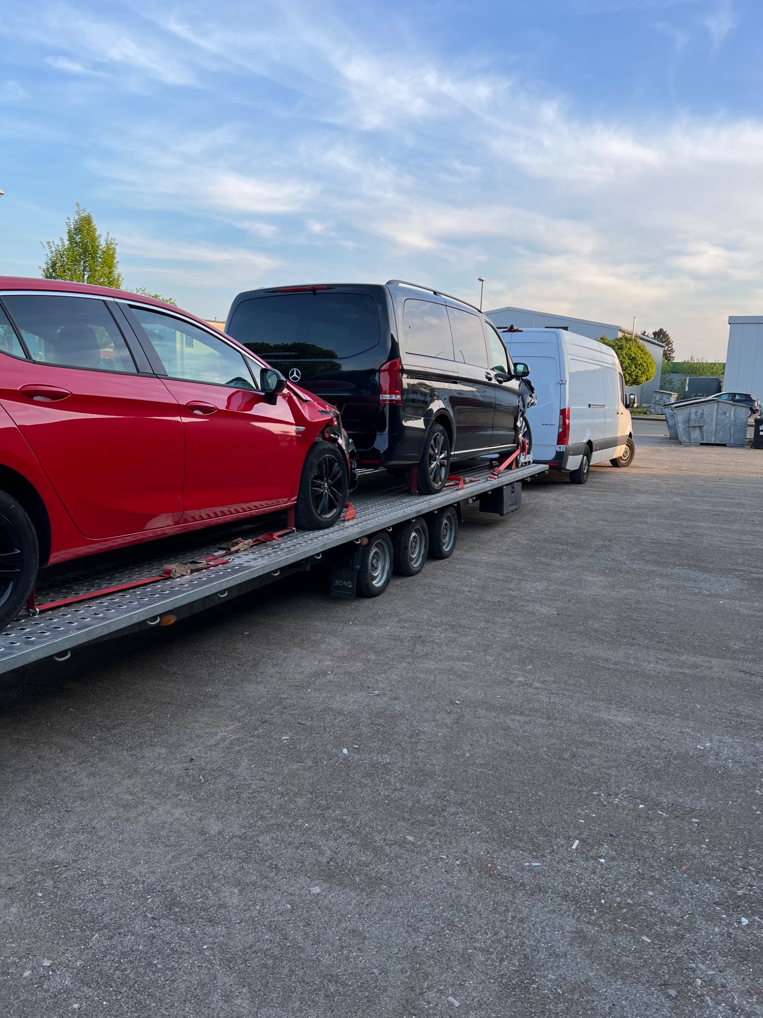 Czerwony samochód z uszkodzonym przodem, czarny van Mercedes i biały bus na lawecie transportowej, zabezpieczone czerwonymi pasami, na tle betonowego placu i błękitnego nieba.
