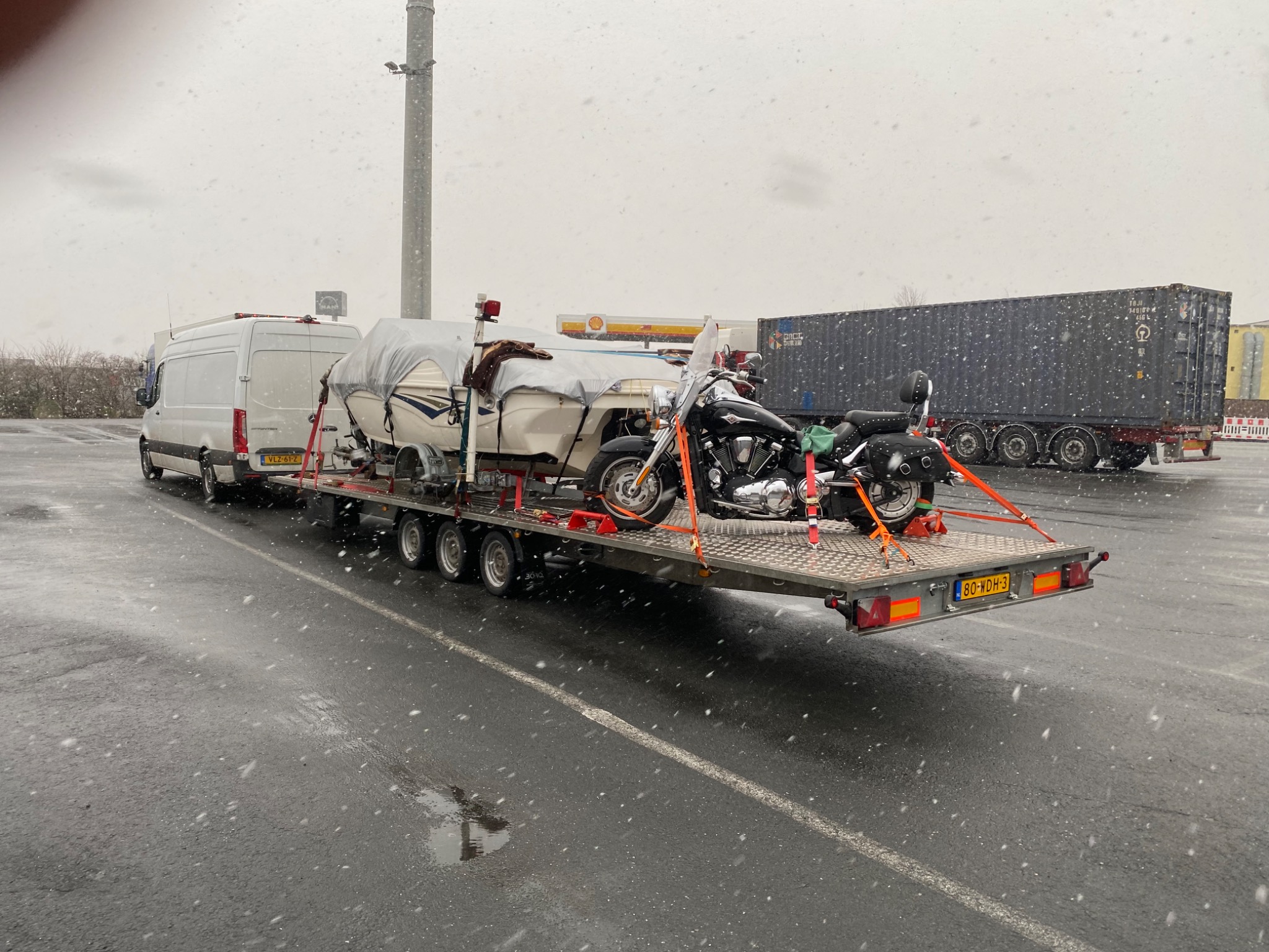 Przyczepa z samochodem dostawczym, przykrytą łodzią i motocyklem, zabezpieczonymi pomarańczowymi pasami transportowymi, na parkingu w zimowej scenerii z padającym śniegiem.