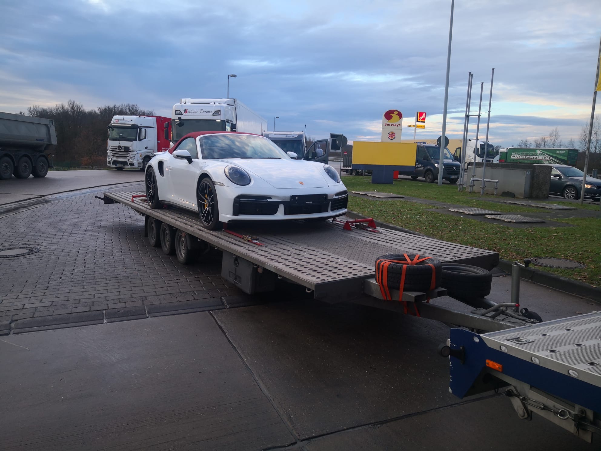 Białe Porsche 911 Cabriolet z czerwonym dachem, zabezpieczone pasami na niskopodwoziowej lawecie samochodowej, na tle stacji benzynowej i zaparkowanych ciężarówek.