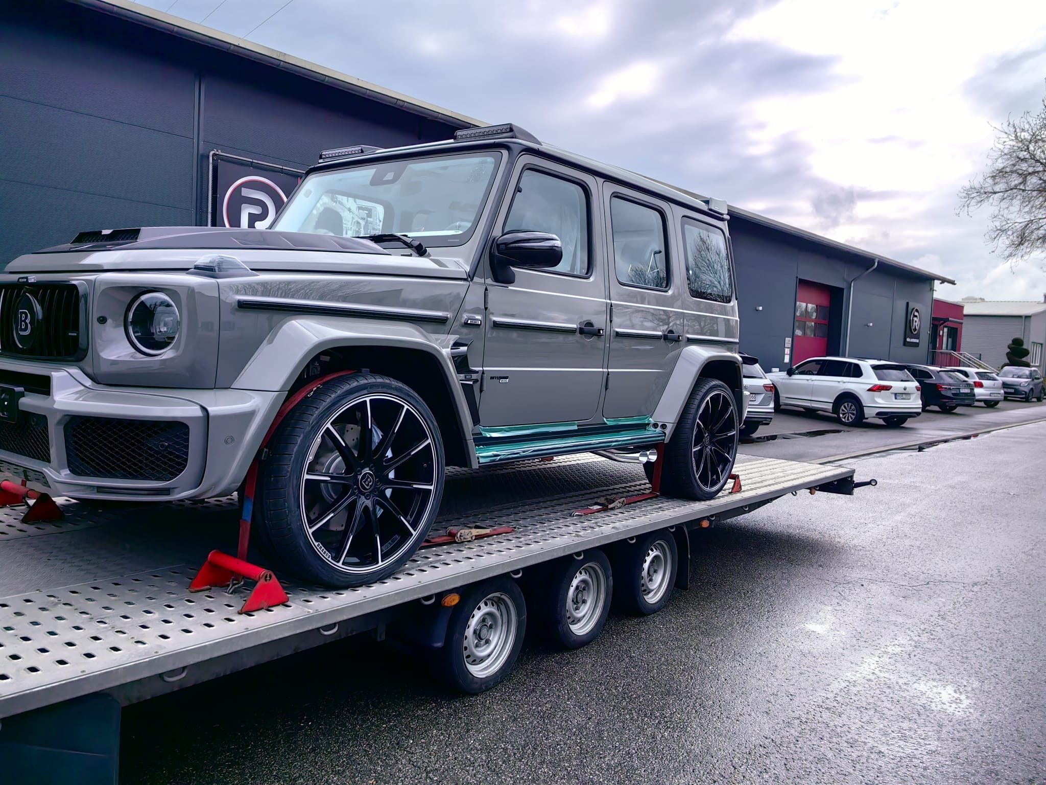 Szary, luksusowy samochód marki Brabus zabezpieczony czerwonymi pasami transportowymi na niskopodwoziowej lawecie, na tle budynku z logo firmy.