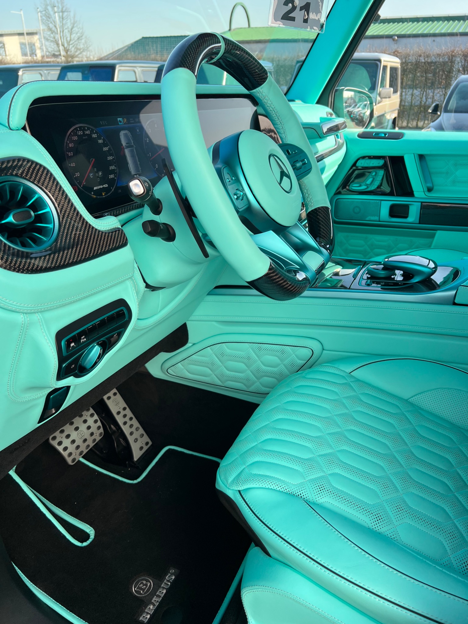 Wnętrze luksusowego samochodu marki Brabus w kolorze miętowym, z detalami z włókna węglowego na kierownicy i desce rozdzielczej, widoczne pedały i dywaniki z logo Brabus.