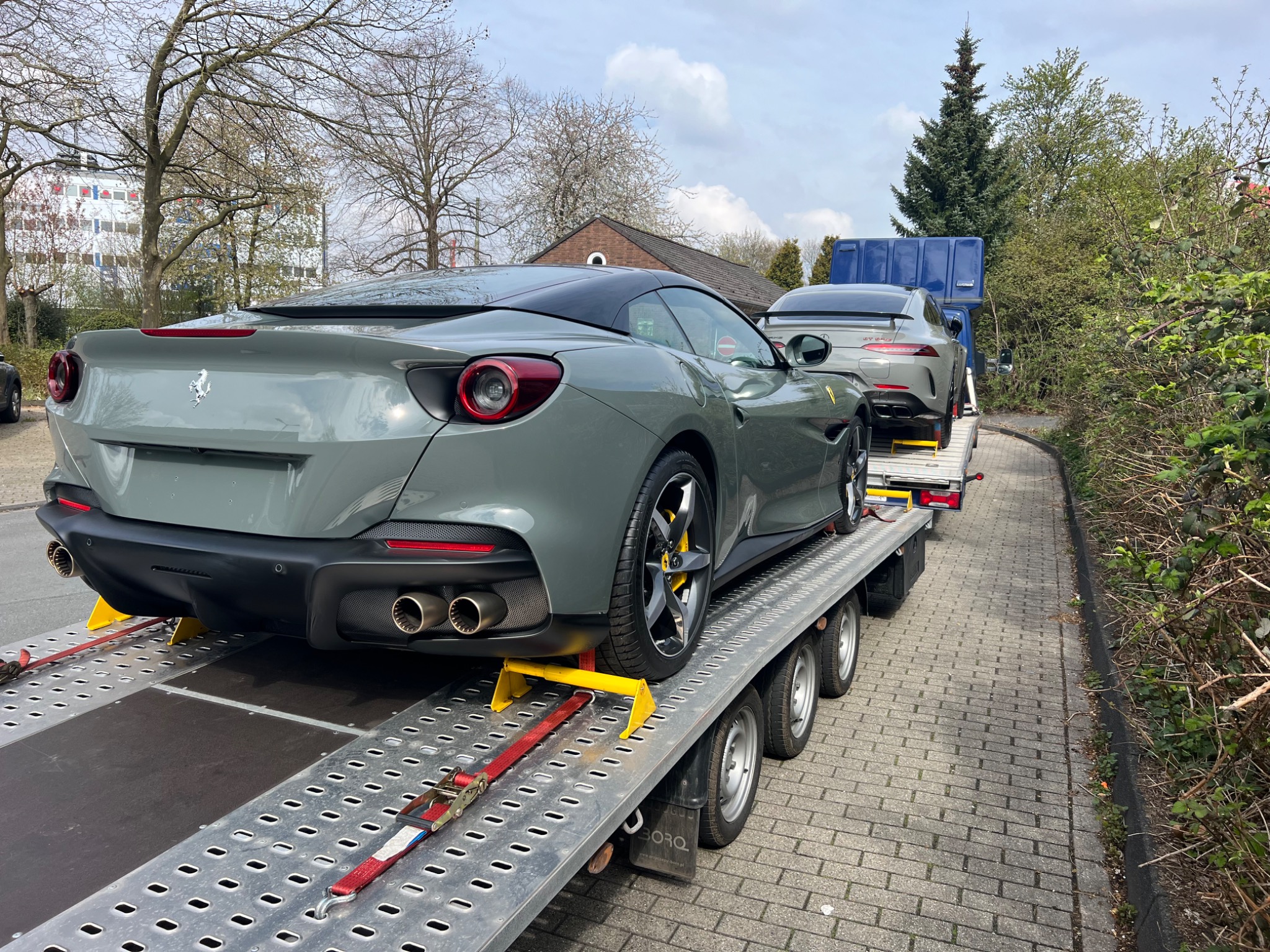 Dwa luksusowe samochody, Ferrari i Mercedes-AMG GT, zabezpieczone pasami na platformie lawety transportowej, widok z tyłu na tle zieleni i zabudowań.