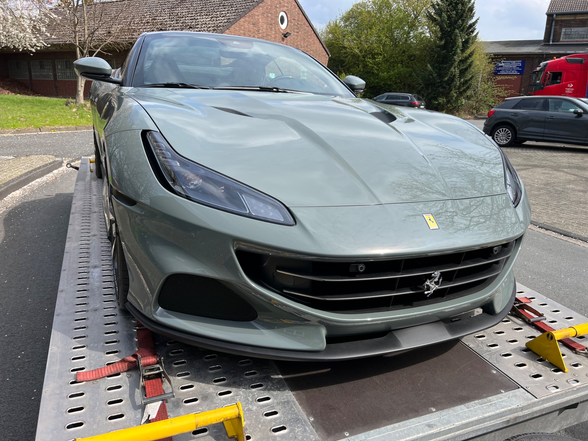 Szary sportowy samochód Ferrari na lawecie, zabezpieczony czerwonymi pasami transportowymi, widok z przodu, w tle budynek i inne pojazdy.