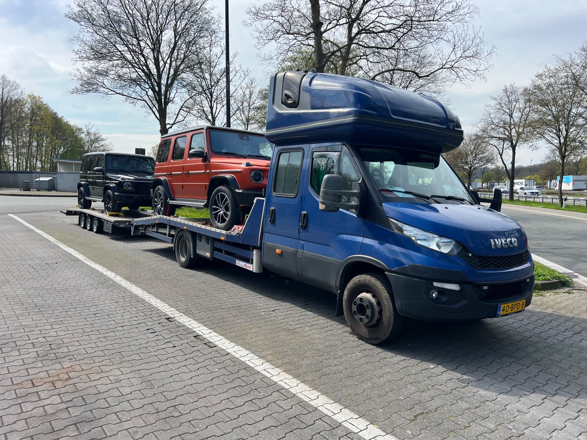 Niebieska laweta z dwoma samochodami Mercedes klasy G (pomarańczowy i czarny) na parkingu w s'-Heerenberg, widok z boku, tablica rejestracyjna widoczna.