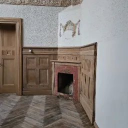 Wnętrze pokoju z parkietem w jodełkę, boazerią, drewnianymi drzwiami i ceglanym kominkiem z paleniskiem. Na ścianie dekoracyjny herb.