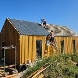 HB BUDOWNICTWO SPÓŁKA Z OGRANICZONĄ ODPOWIEDZIALNOŚCIĄ - Budowa domu z drewnianą elewacją: dwaj mężczyźni montują szarą blachodachówkę na dachu, widoczne rusztowanie i elementy izolacji fundamentów, w tle krajobraz polny i błękitne niebo.