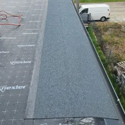 Fragment dachu płaskiego z papą termozgrzewalną Nexler, widok z góry, etap prac dekarskich. W tle budynki mieszkalne i biały samochód dostawczy.