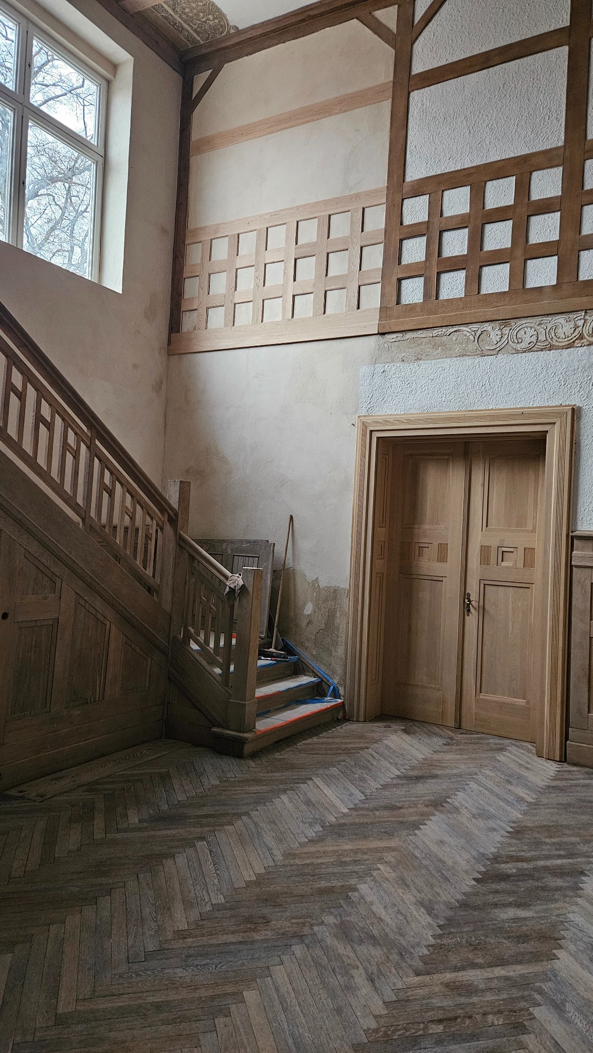 Wnętrze domu w trakcie remontu: drewniane schody zabezpieczone folią, parkiet ułożony w jodełkę, surowe drewniane elementy dekoracyjne na ścianach i nowe drewniane drzwi.