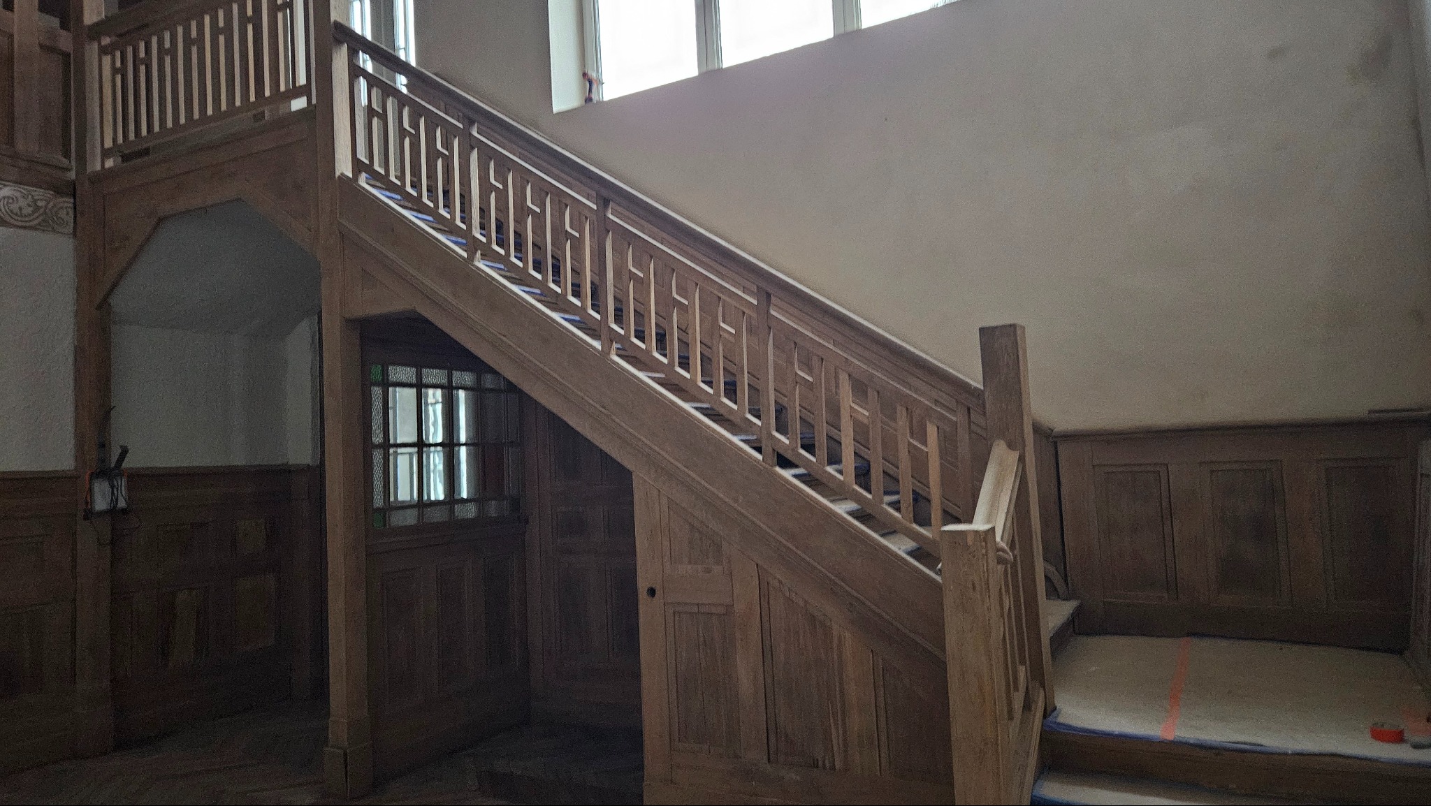 Drewniane schody z balustradą w odnawianym wnętrzu domu, zabezpieczone folią malarską, widoczne okno i panele ścienne.