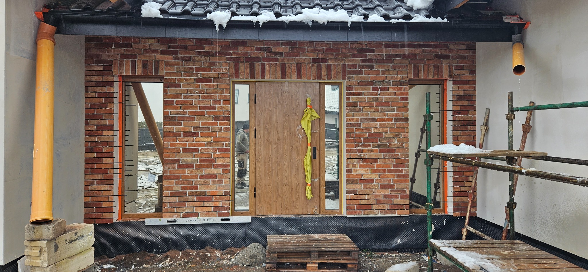 Fragment ściany zewnętrznej domu z nową, częściowo wykonaną elewacją z cegły klinkierowej, widoczne rusztowania, orynnowanie i folia hydroizolacyjna przy fundamentach.