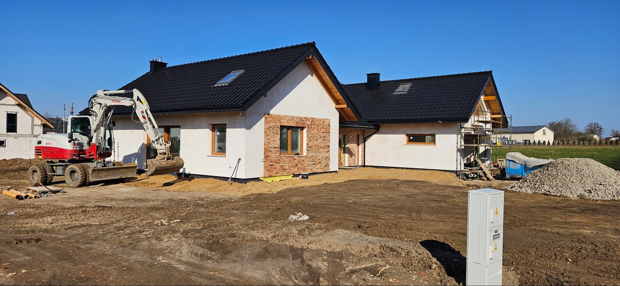 Nowoczesne domy jednorodzinne w trakcie budowy, z częściowo wykończoną elewacją z cegły klinkierowej, widoczny mały biało-czerwony koparko-ładowarka na tle błękitnego nieba.