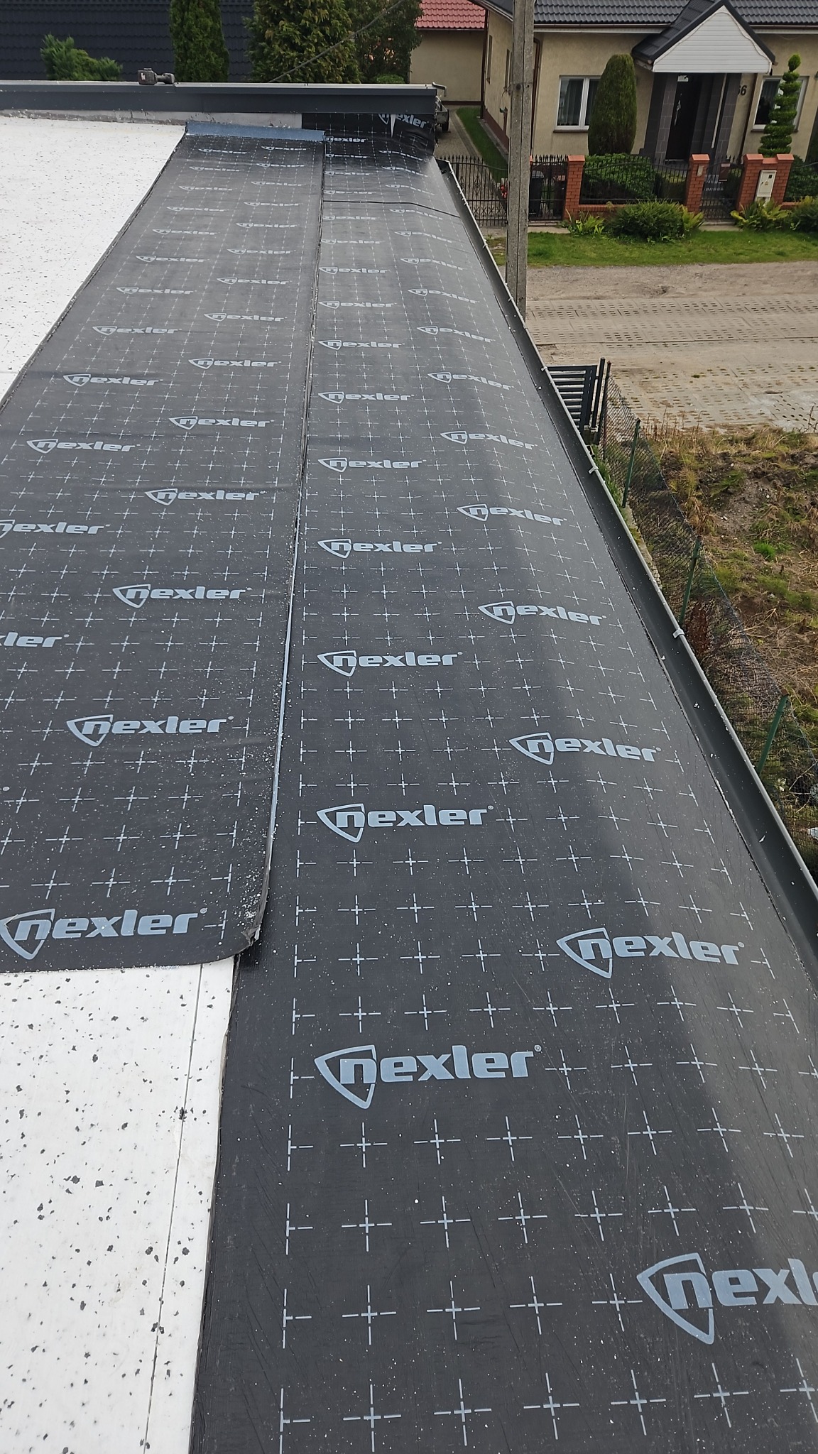 Czarny materiał izolacyjny z logo Nexler, częściowo rozłożony na białej powierzchni dachu płaskiego, widok z góry, z fragmentem budynku w tle.