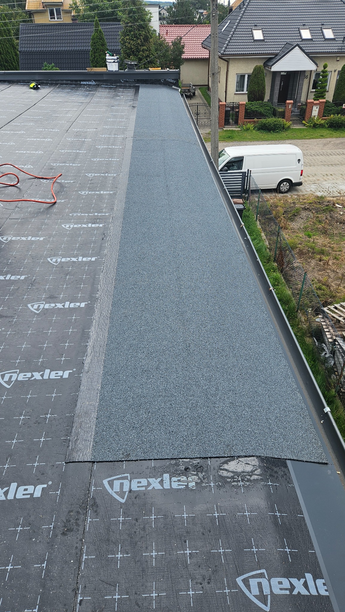 Fragment dachu płaskiego z papą termozgrzewalną Nexler, widok z góry, etap prac dekarskich. W tle budynki mieszkalne i biały samochód dostawczy.