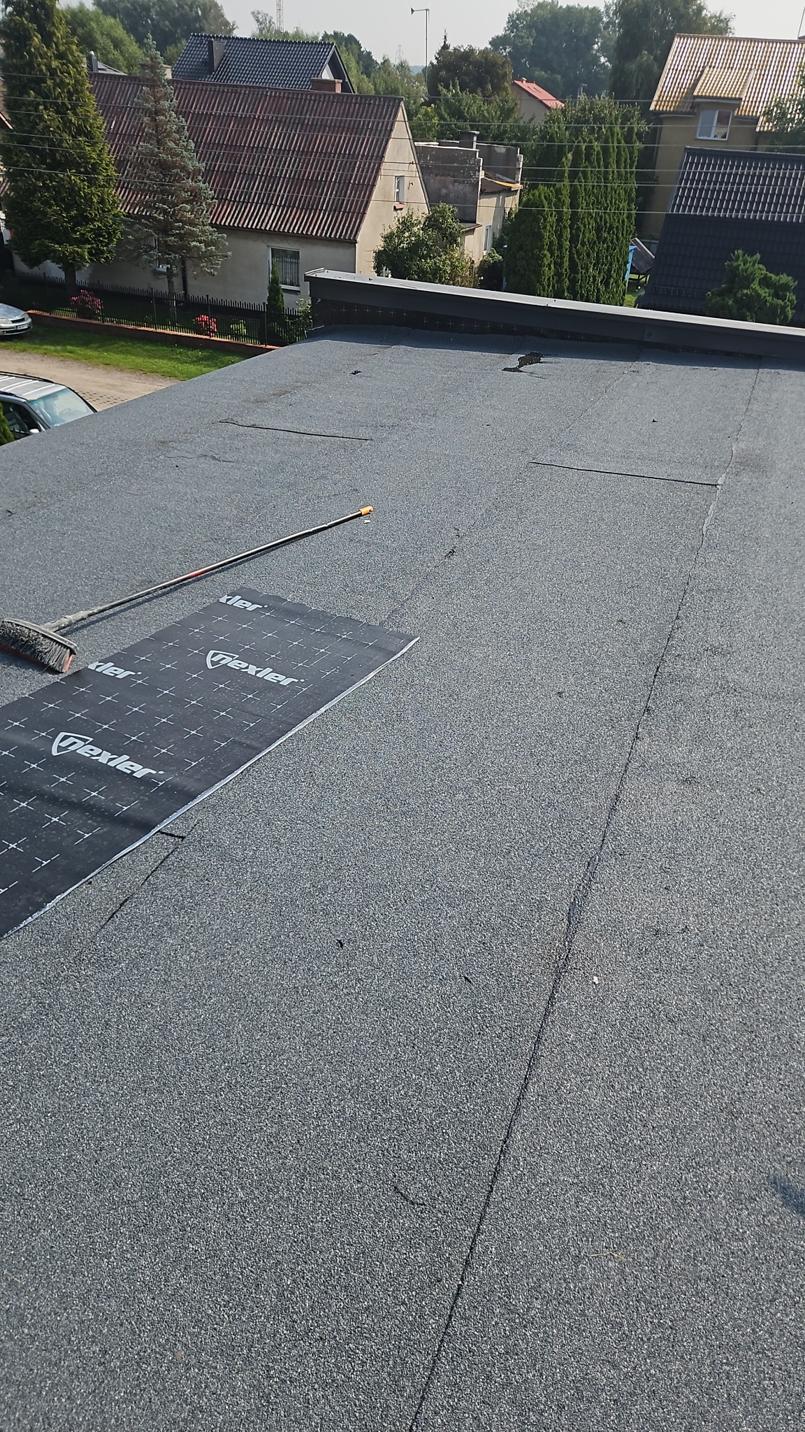 Fragment dachu pokrytego papą z widocznym arkuszem papy i szczotką. W tle zabudowa jednorodzinna.