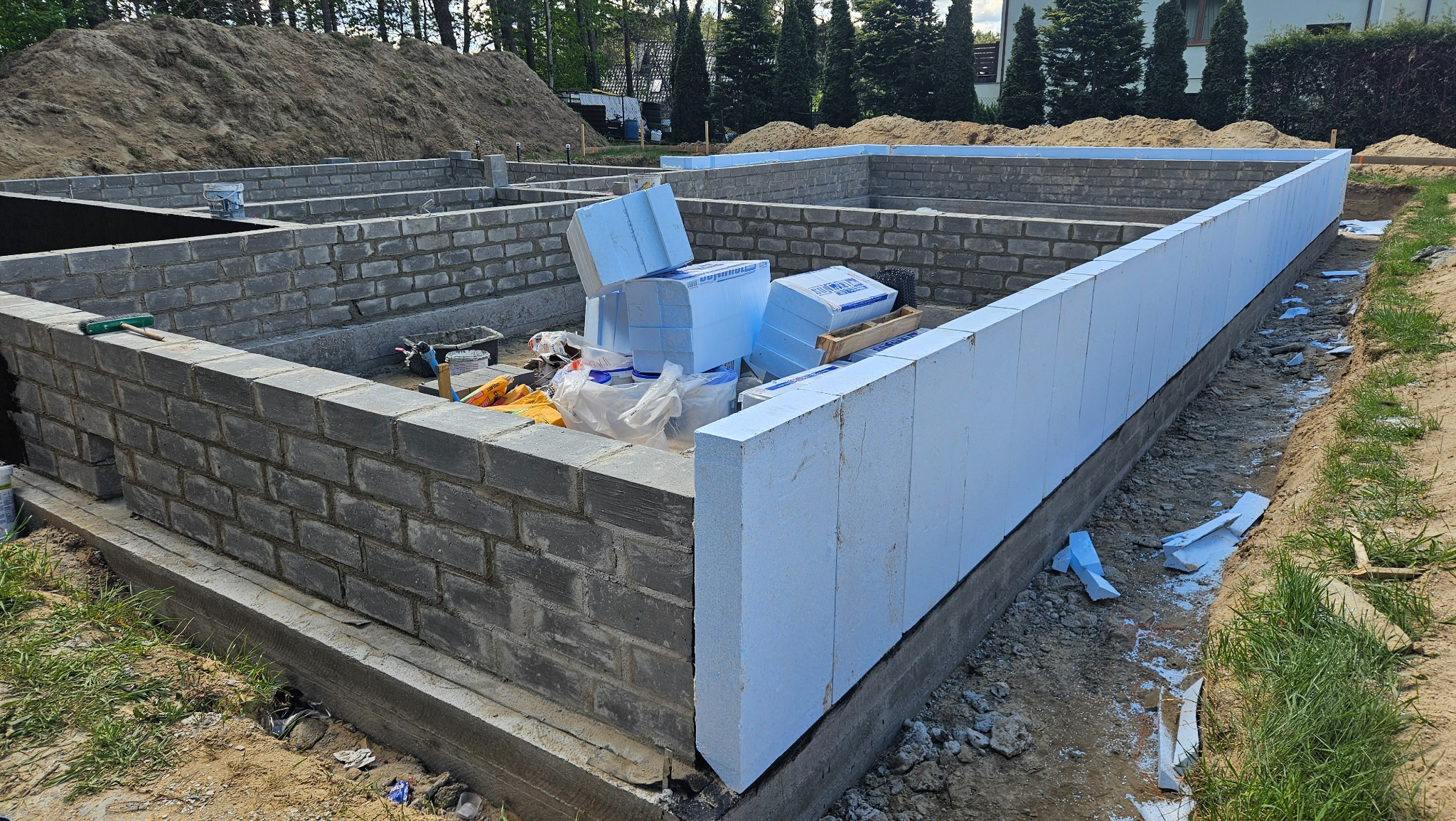 Fundamenty domu w trakcie budowy: ściany z szarej cegły i ocieplenie z niebieskiego styropianu. Na fundamencie leżą materiały budowlane.