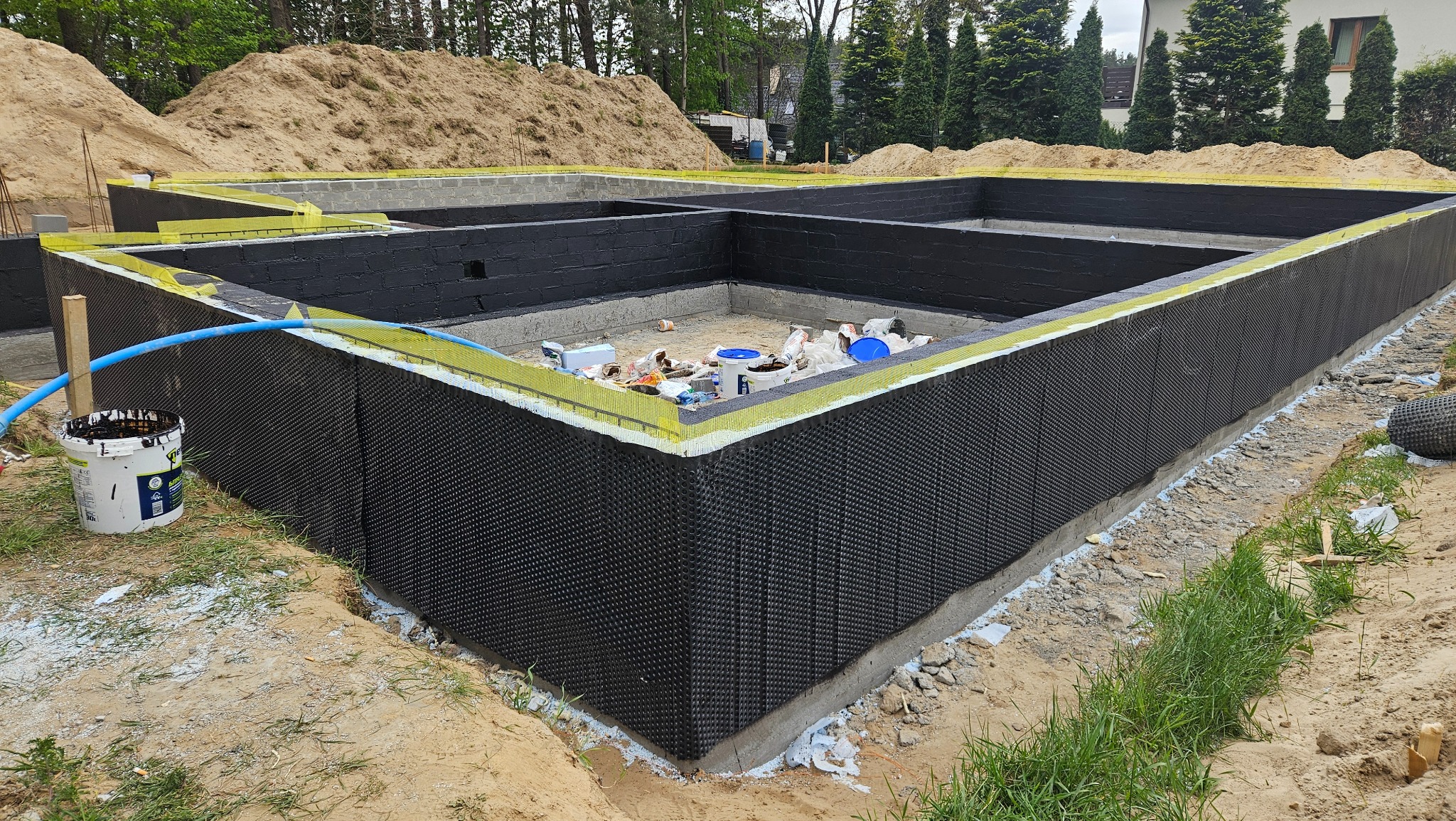 Izolacja fundamentów budynku z użyciem folii kubełkowej i pianki, widoczne bloczki fundamentowe i zasypka ziemna w tle. Etap prac budowlanych.