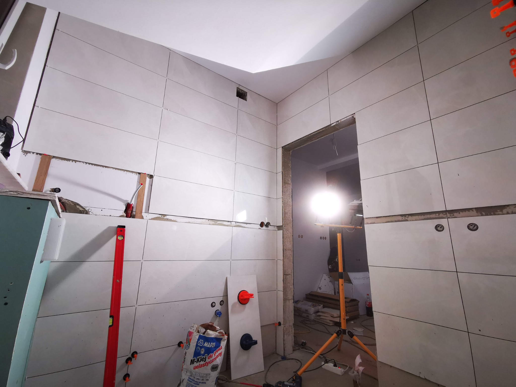 Układanie białych, prostokątnych płytek na ścianie w trakcie remontu łazienki, widoczny poziomica, przyssawki do płytek, worek z klejem i lampa budowlana na trójnogu.