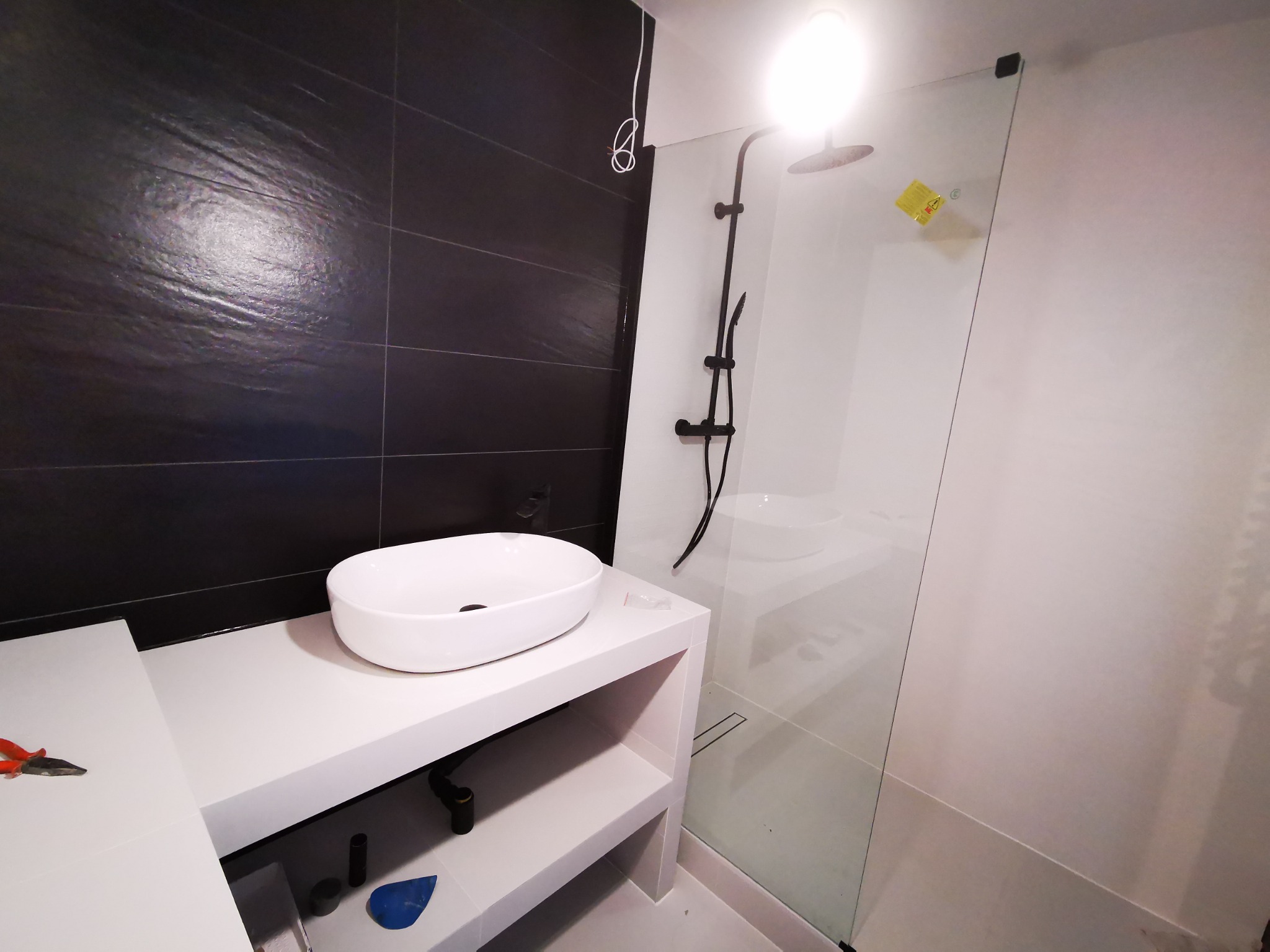 Nowoczesna łazienka z czarnymi płytkami na ścianie, białą umywalką nablatową i kabiną prysznicową ze szklaną ścianką, widoczne narzędzia glazurnicze.