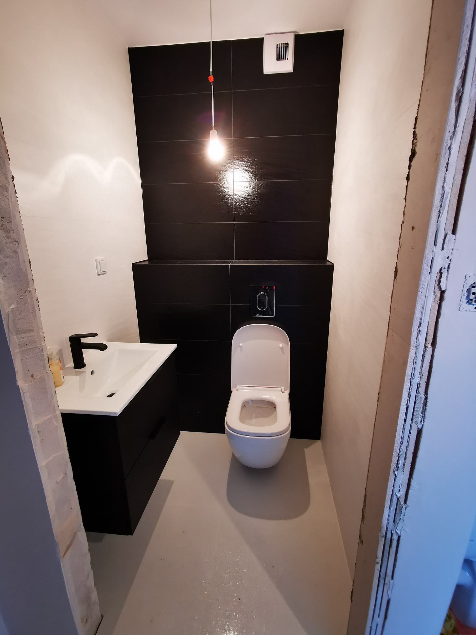 Nowoczesna łazienka w trakcie remontu: czarne płytki na ścianie, biała umywalka z czarną armaturą, wisząca toaleta i surowa ściana z widocznym profilem.