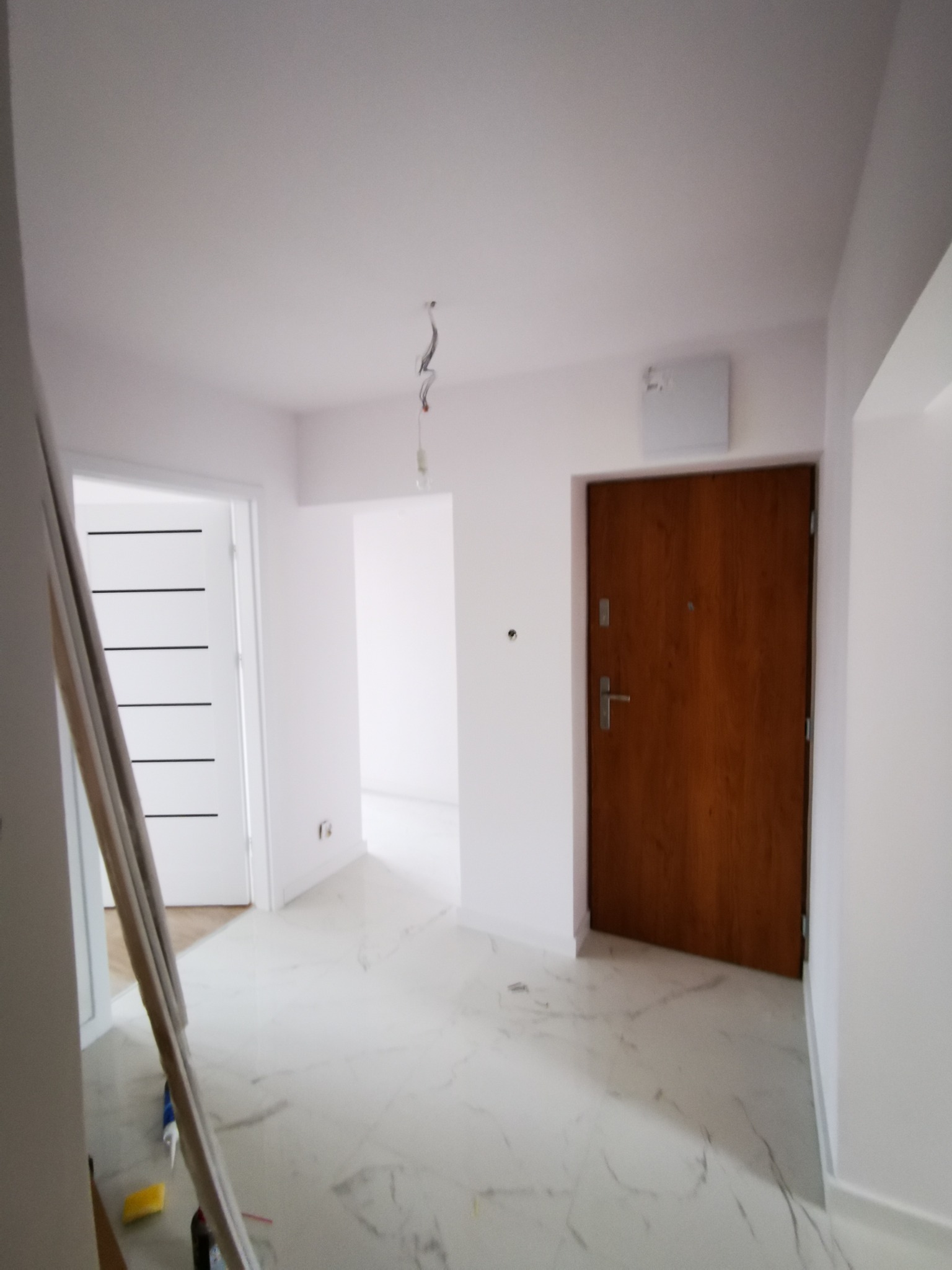 Wnętrze po remoncie: białe ściany, podłoga imitująca marmur, drewniane drzwi wejściowe i białe drzwi wewnętrzne z czarnymi paskami. Na podłodze leżą listwy przypodłogowe i narzędzia.