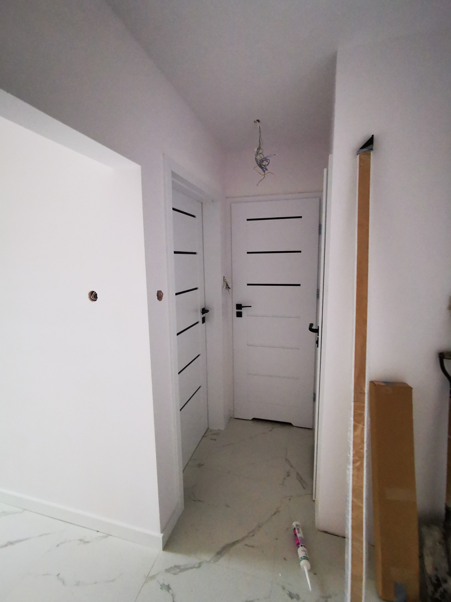Korytarz w trakcie prac wykończeniowych, widoczne dwie białe drzwi z czarnymi poziomymi paskami, białe ściany z otworami po gniazdkach, marmurowe płytki podłogowe oraz materiały budowlane.