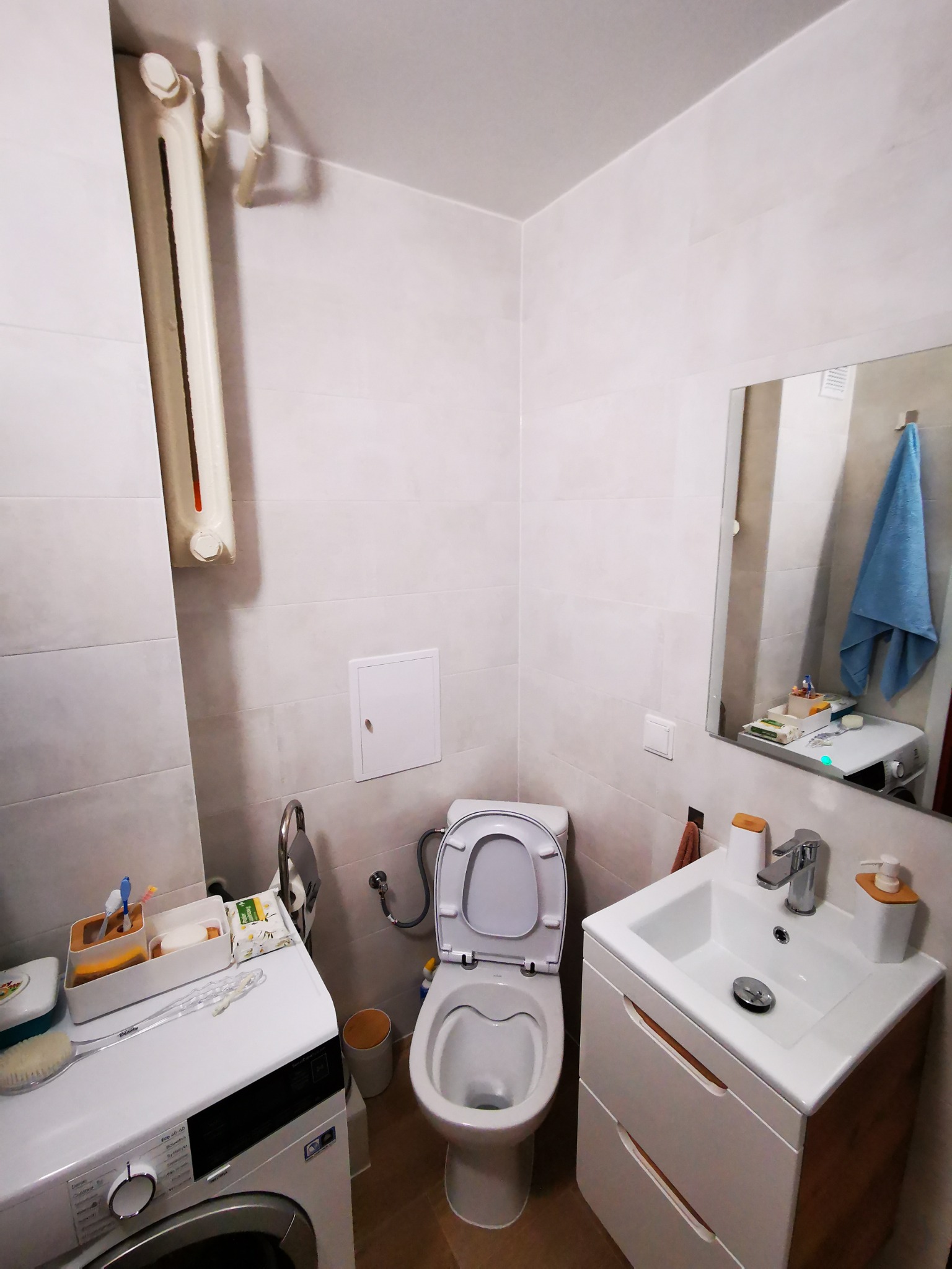 Jasna łazienka z toaletą z otwartą klapą, umywalką z szafką z drewnianymi elementami oraz pralką z akcesoriami na górze, widok na kafelkowane ściany i grzejnik.