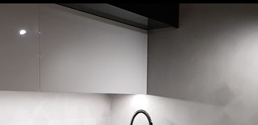 Fragment nowoczesnej kuchni z szarymi, lakierowanymi szafkami wiszącymi i czarnym okapem, oświetlenie punktowe pod szafkami, widoczny fragment baterii kuchennej.