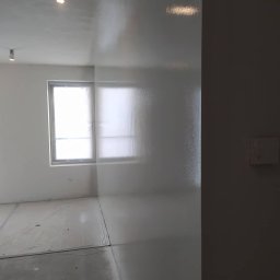 Firma MALOREM - Świeżo wykończone, gładkie, białe ściany w pokoju z oknem zabezpieczonym folią, podłoga również zabezpieczona, widoczne delikatne refleksy światła.