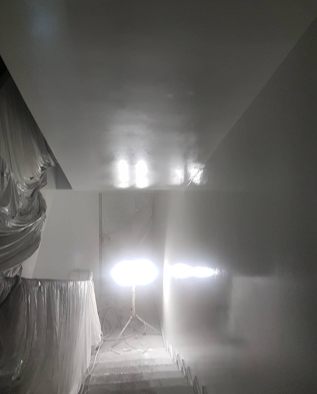 Świeżo pokryte białą farbą ściany klatki schodowej, zabezpieczone folią meble i podłoga, oświetlone lampą budowlaną na trójnogu.