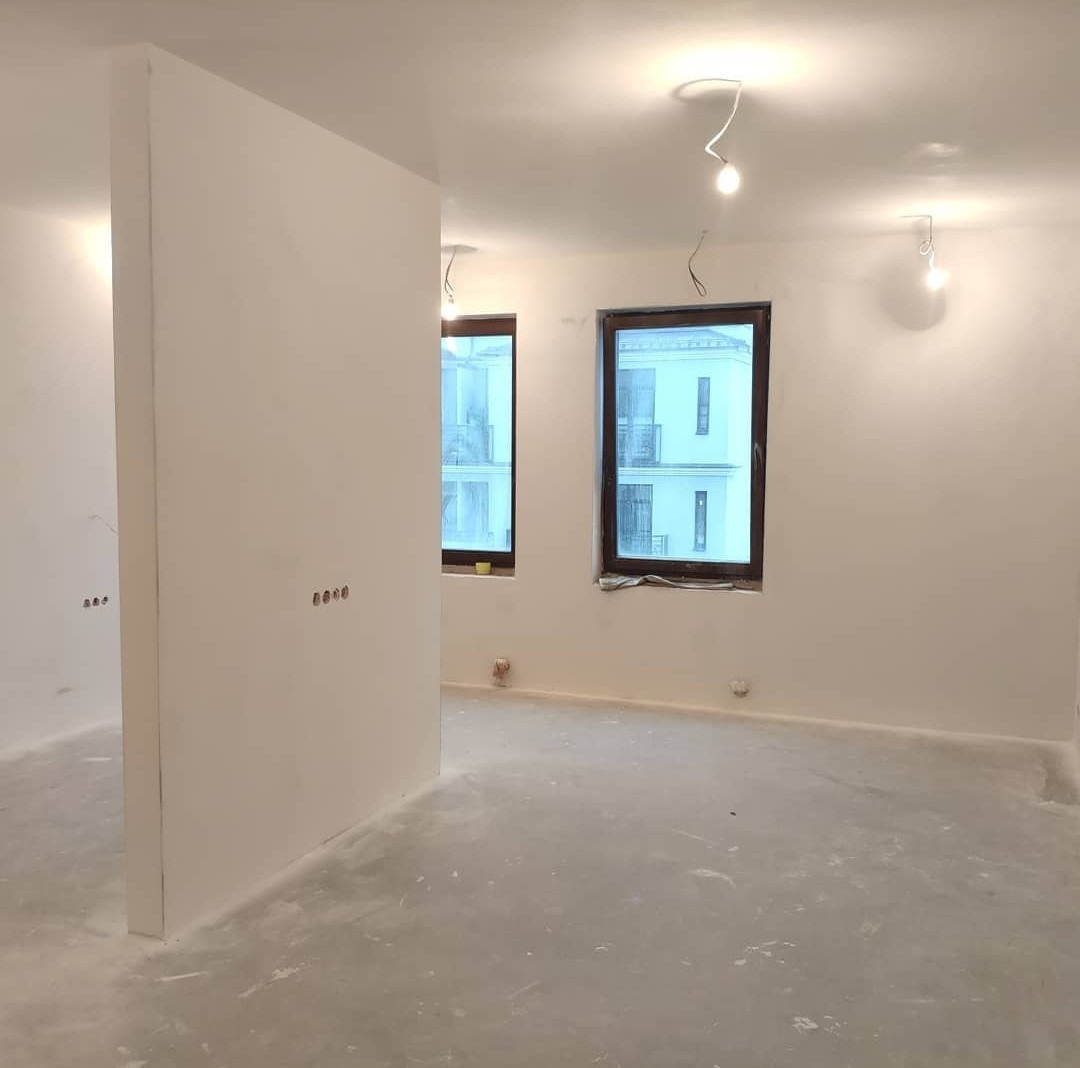 Wnętrze w trakcie przygotowania do wykończenia, widoczne świeżo wygładzone ściany, surowa podłoga i okna z widokiem na budynek mieszkalny.