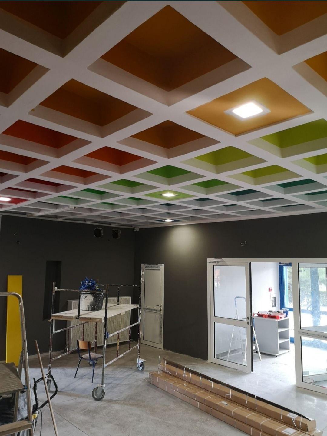 Wielobarwny kasetonowy sufit w odcieniach pomarańczy, zieleni i czerwieni, widoczny z poziomu podłogi z rusztowaniem i materiałami budowlanymi w pomieszczeniu biurowym.