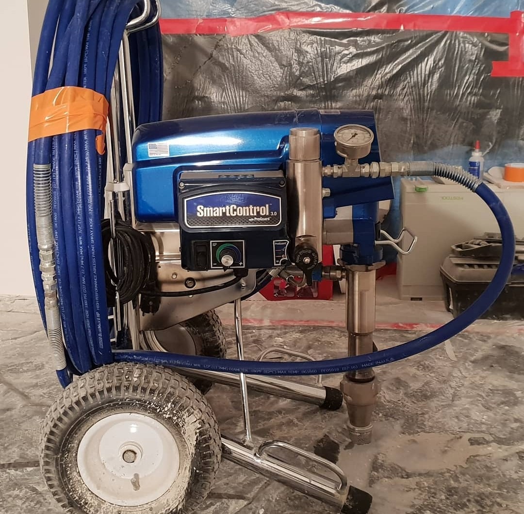 Profesjonalny, niebieski agregat malarski SmartControl 3.0 na kółkach, z widocznymi przewodami, manometrem i osłoną podłogi z folii, przygotowany do prac wykończeniowych.