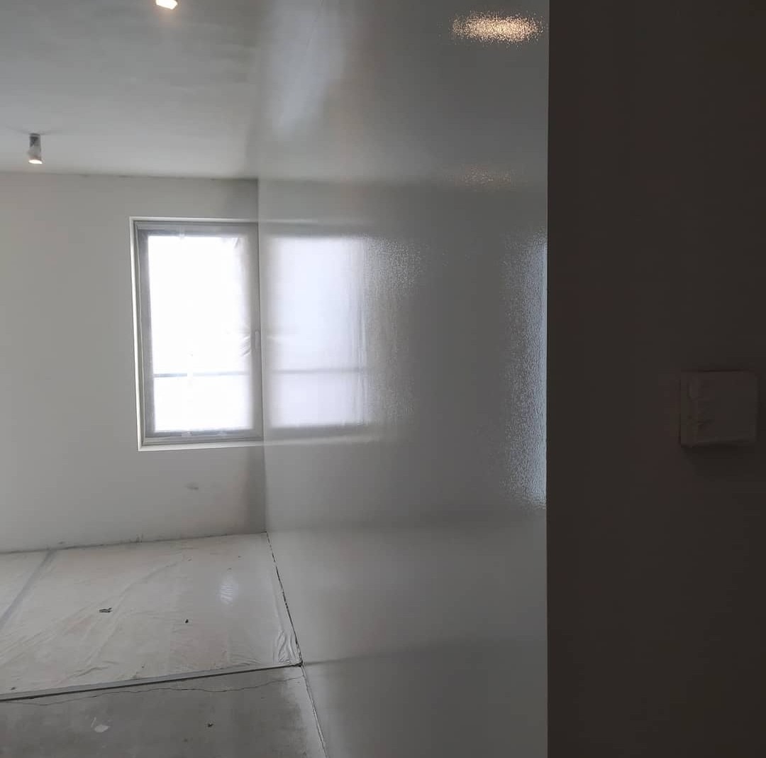 Świeżo wykończone, gładkie, białe ściany w pokoju z oknem zabezpieczonym folią, podłoga również zabezpieczona, widoczne delikatne refleksy światła.