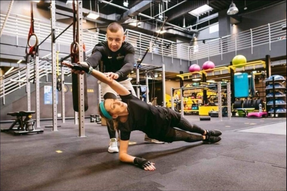 Kobieta wykonuje ćwiczenie deski bokiem z wyciągniętą ręką, pod nadzorem trenera personalnego w nowoczesnej siłowni z widocznymi drabinkami, piłkami gimnastycznymi i innym sprzętem.