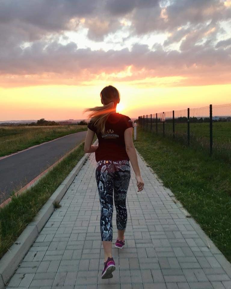 Kobieta w sportowych legginsach idzie ścieżką w kierunku zachodzącego słońca, widok z tyłu.