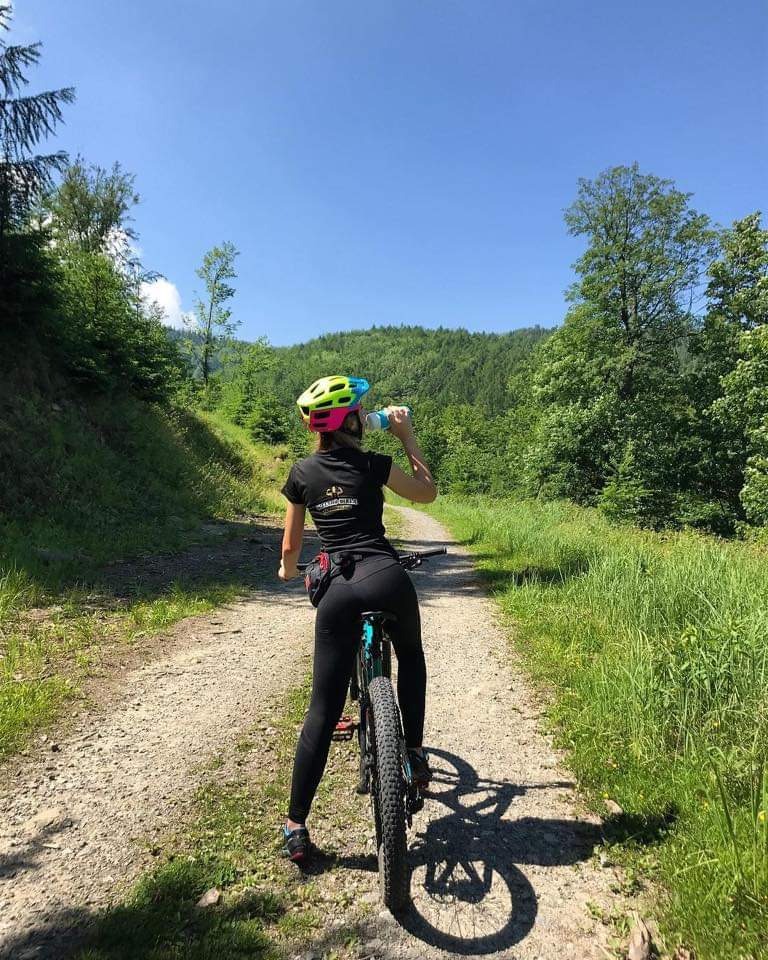 Kobieta w czarnym stroju sportowym i kasku pije wodę podczas jazdy na rowerze górskim po polnej drodze w lesie, widok z tyłu.