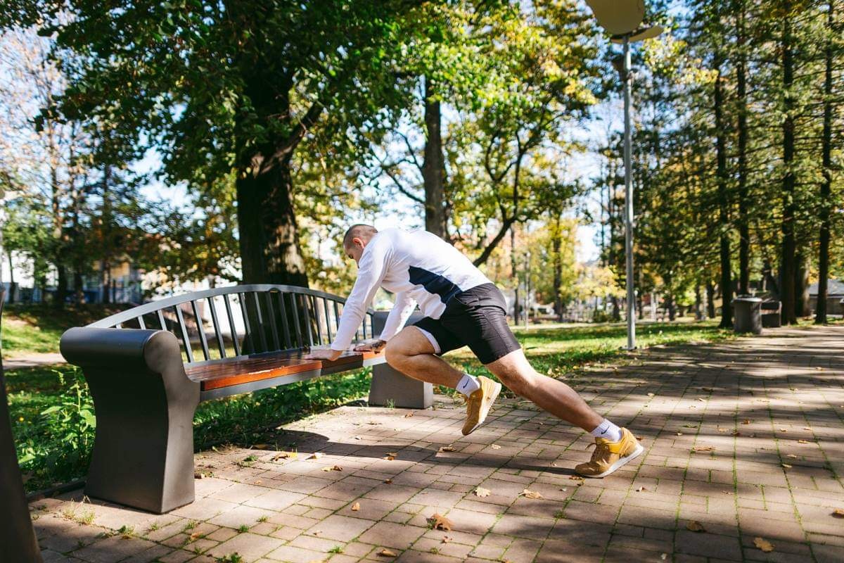Mężczyzna w sportowym stroju wykonuje ćwiczenie rozciągające na ławce w parku, w tle drzewa i ścieżka z kostki brukowej.