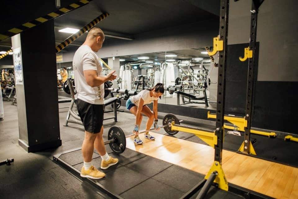 Kobieta w białej koszulce i niebieskich spodenkach podnosi sztangę pod okiem mężczyzny w żółtych butach w siłowni z lustrami i sprzętem do ćwiczeń.
