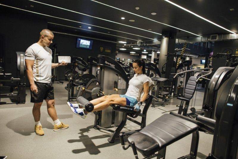 Kobieta ćwiczy na maszynie do nóg w siłowni, nadzorowana przez mężczyznę z tabletem w ręku, na tle innych maszyn i czarnego sufitu z białymi liniami świetlnymi.