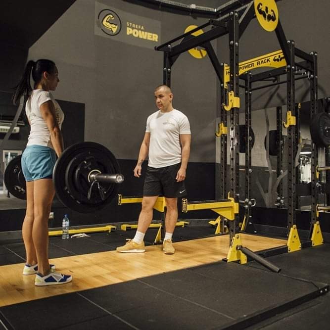 Kobieta w trakcie podnoszenia sztangi pod okiem mężczyzny w siłowni z logiem 'Strefa Power' w tle.