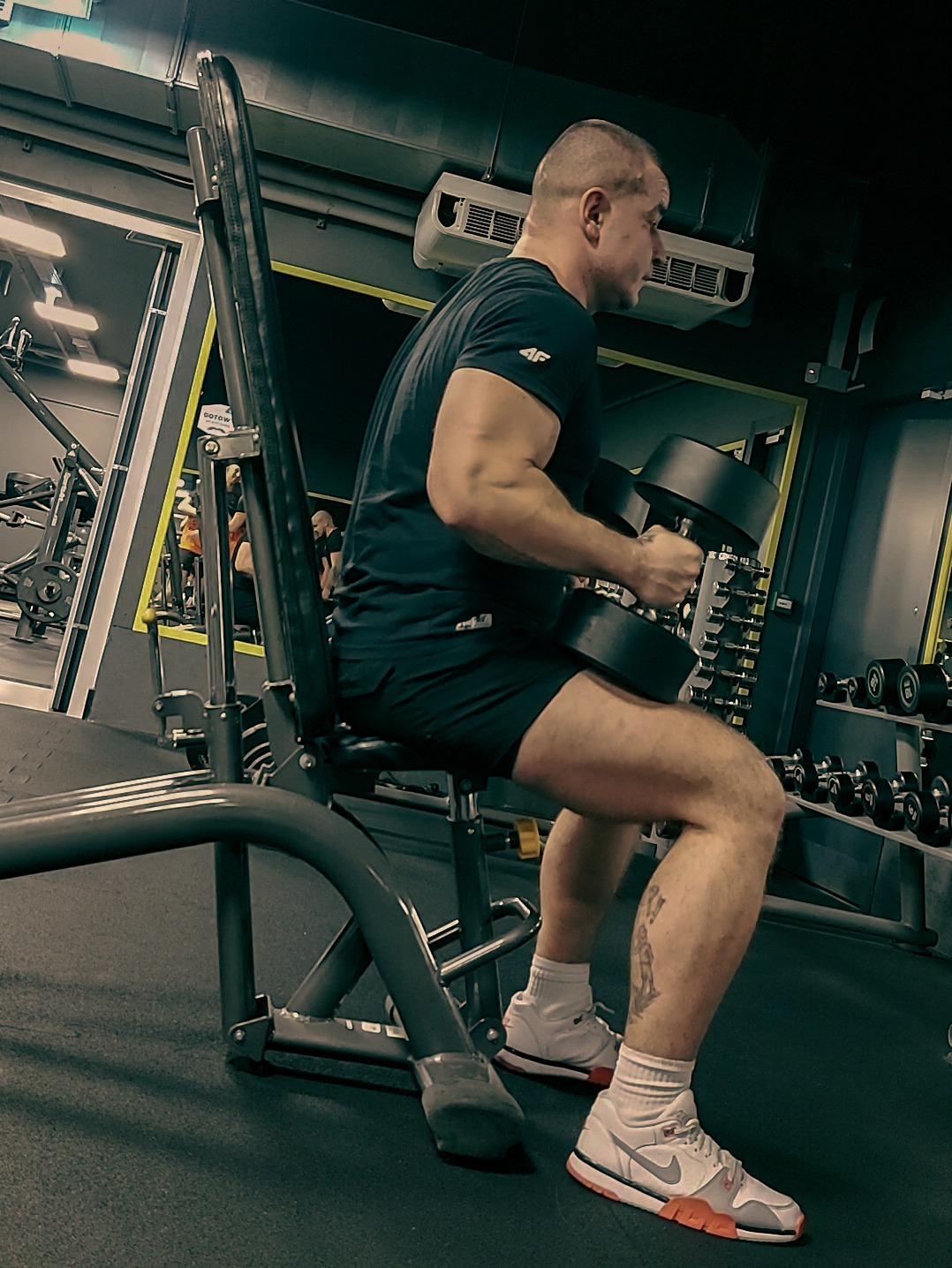 Mężczyzna w czarnej koszulce i spodenkach siedzi na ławce w siłowni, trzymając hantle. Widoczny tatuaż na łydce i białe buty sportowe.
