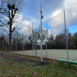 Eye'sCAM - Kamera monitoringu na słupie obok boiska z koszem, otoczona siatką. Widok na zewnątrz, jesienne liście na ziemi, pochmurne niebo. Bez osób.