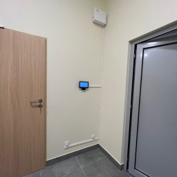 Eye'sCAM - Panel sterowania systemem alarmowym zamontowany na ścianie wewnątrz budynku. Widoczne przewody w listwach, drzwi wejściowe i fragment podłogi z szarych płytek.