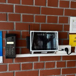 Eye'sCAM - Zamontowany wideodomofon SLICAN z panelem dotykowym i klawiaturą numeryczną na ceglanej ścianie w szkole, obok tabliczka z prośbą o dzwonienie.