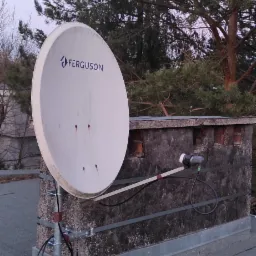 Antena satelitarna Ferguson zamontowana na kominie z widocznymi śladami wilgoci, na dachu, z kablem i śrubokrętem obok.