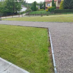 Widok na zadbany ogród z równo przystrzyżonym trawnikiem, żwirową ścieżką i obrzeżami, w tle domy i starannie przycięte tuje.